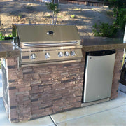 Outdoor kitchen with stainless steel grill and refrigerator against a stone wall.