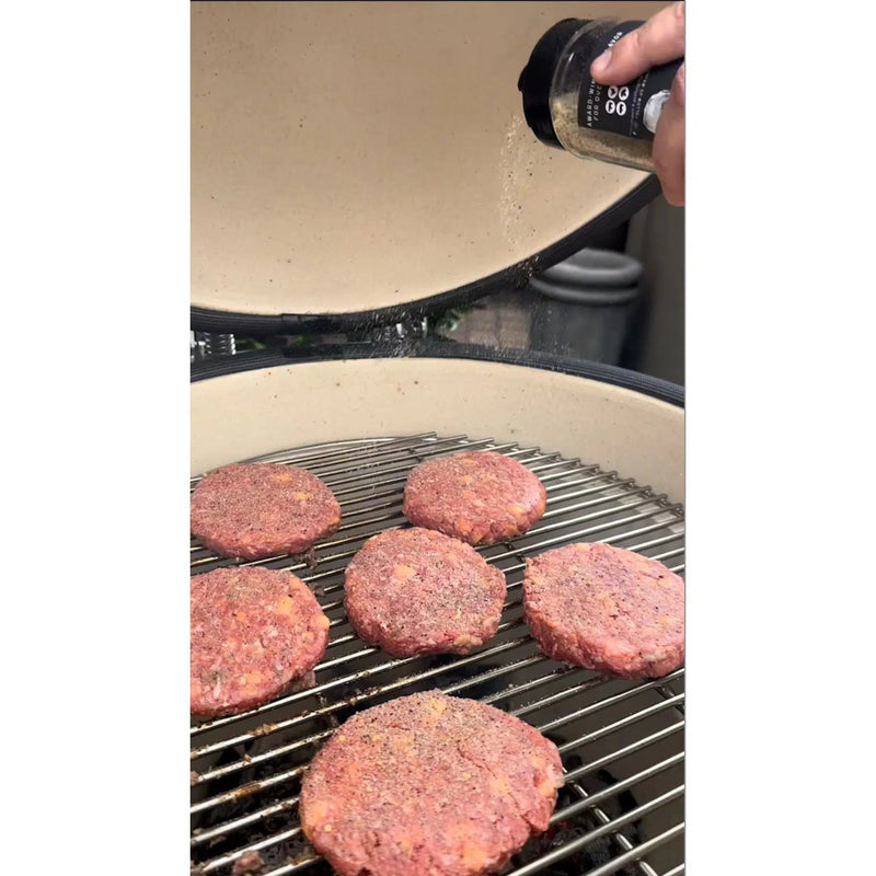 Raw burger patties on a grill with a hand holding a seasoning shaker.