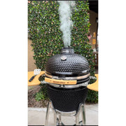 Black kamado-style grill with smoke rising, set against a green hedge.